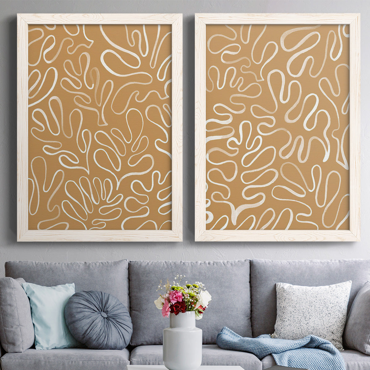 Set of two white barnwood framed canvas prints depicting paired graphic botanical pattern abstracts with leaf-like contours, flat warm backgrounds, and repeated flowing shapes above a sofa in a contemporary living room.