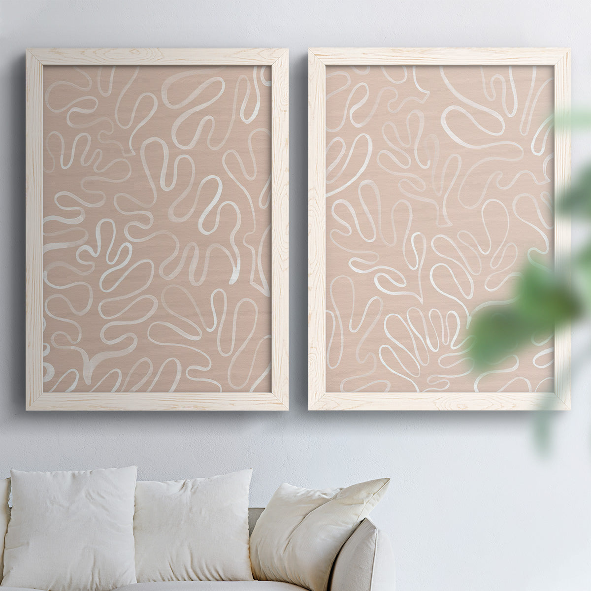 Set of two white barnwood framed canvas prints depicting leaf-like contours, flat warm backgrounds, and repeated flowing shapes arranged across two complementary compositions above a sofa in a bright living space.
