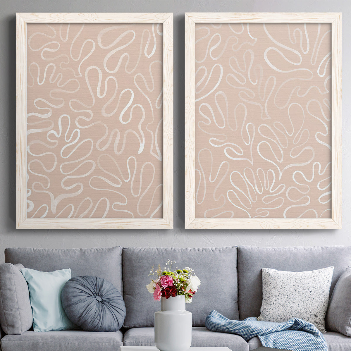 Set of two white barnwood framed canvas prints depicting paired graphic botanical pattern abstracts with leaf-like contours, flat warm backgrounds, and repeated flowing shapes above a sofa in a contemporary living room.