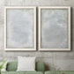 Set of two white barnwood framed canvas prints depicting a coordinated art pair of layered white and silver-gray painted surfaces rendered as quiet textural abstracts above a sofa in a modern living room.