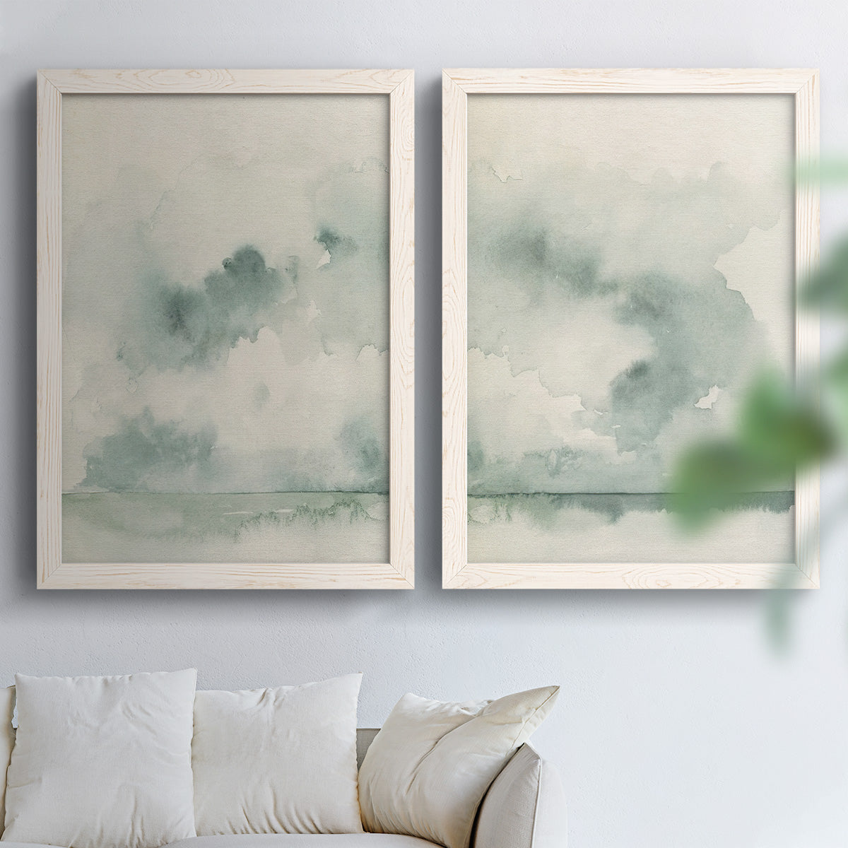 Set of two white barnwood framed canvas prints depicting abstract ocean horizons rendered in translucent gray green watercolor and quiet atmospheric layers above a sofa in a bright living space.