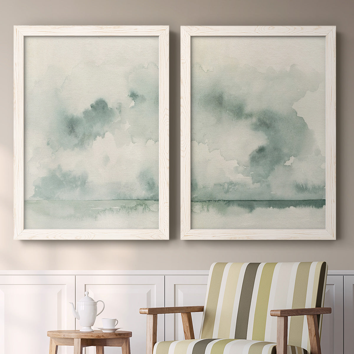 Set of two white barnwood framed canvas prints depicting two minimalist watercolor seascapes with misty green clouds calm horizons and pale reflective water above a sideboard in a cozy sitting room.