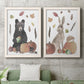 Set of two white barnwood framed canvas prints depicting two whimsical autumn illustrations of forest animals gathered among pumpkins apples feathers and seasonal foliage above a sofa in a modern living room.