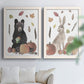 Set of two white barnwood framed canvas prints depicting a rustic harvest set featuring a black bear gray rabbit hedgehog fruit and scattered leaves above a sofa in a bright living space.