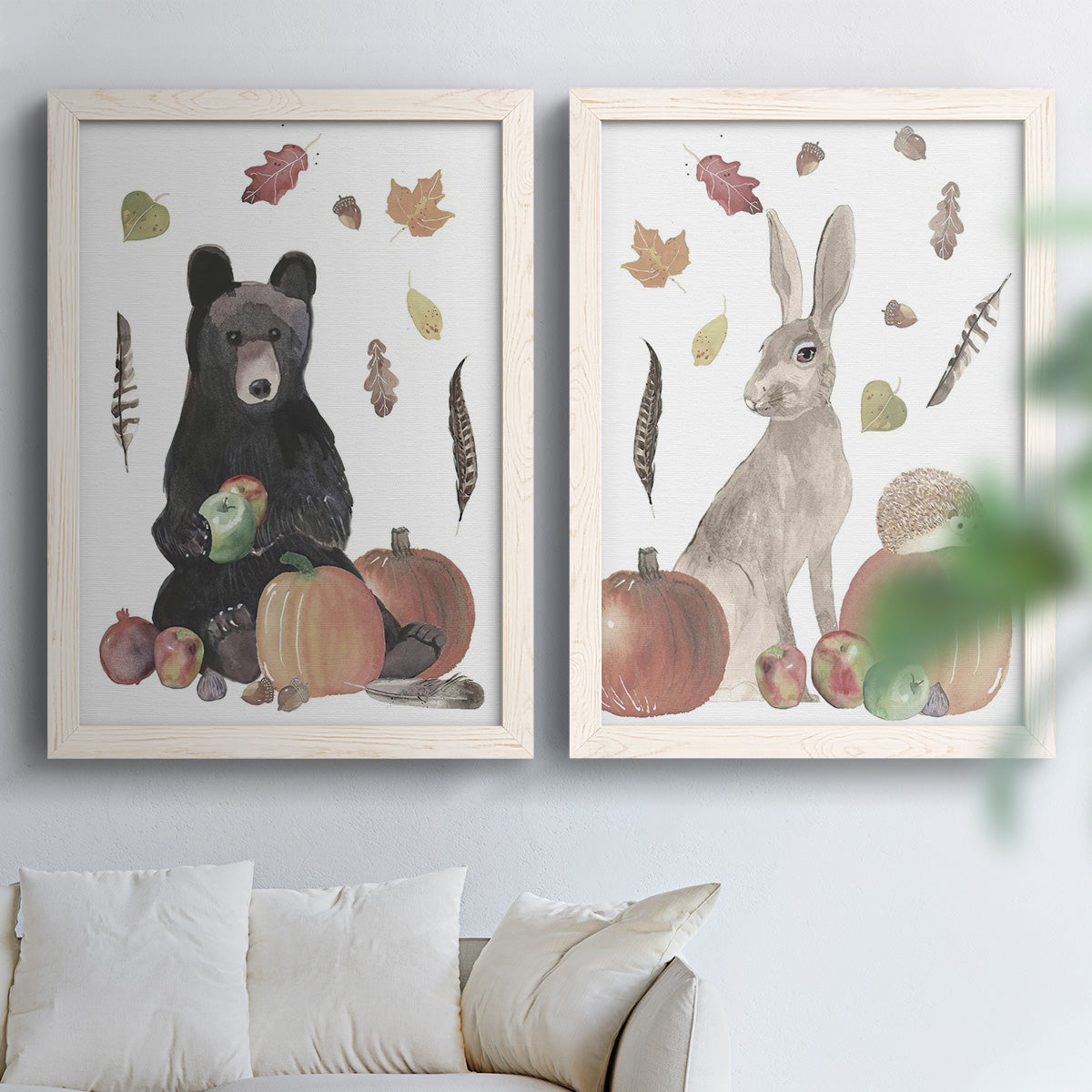 Set of two white barnwood framed canvas prints depicting a rustic harvest set featuring a black bear gray rabbit hedgehog fruit and scattered leaves above a sofa in a bright living space.