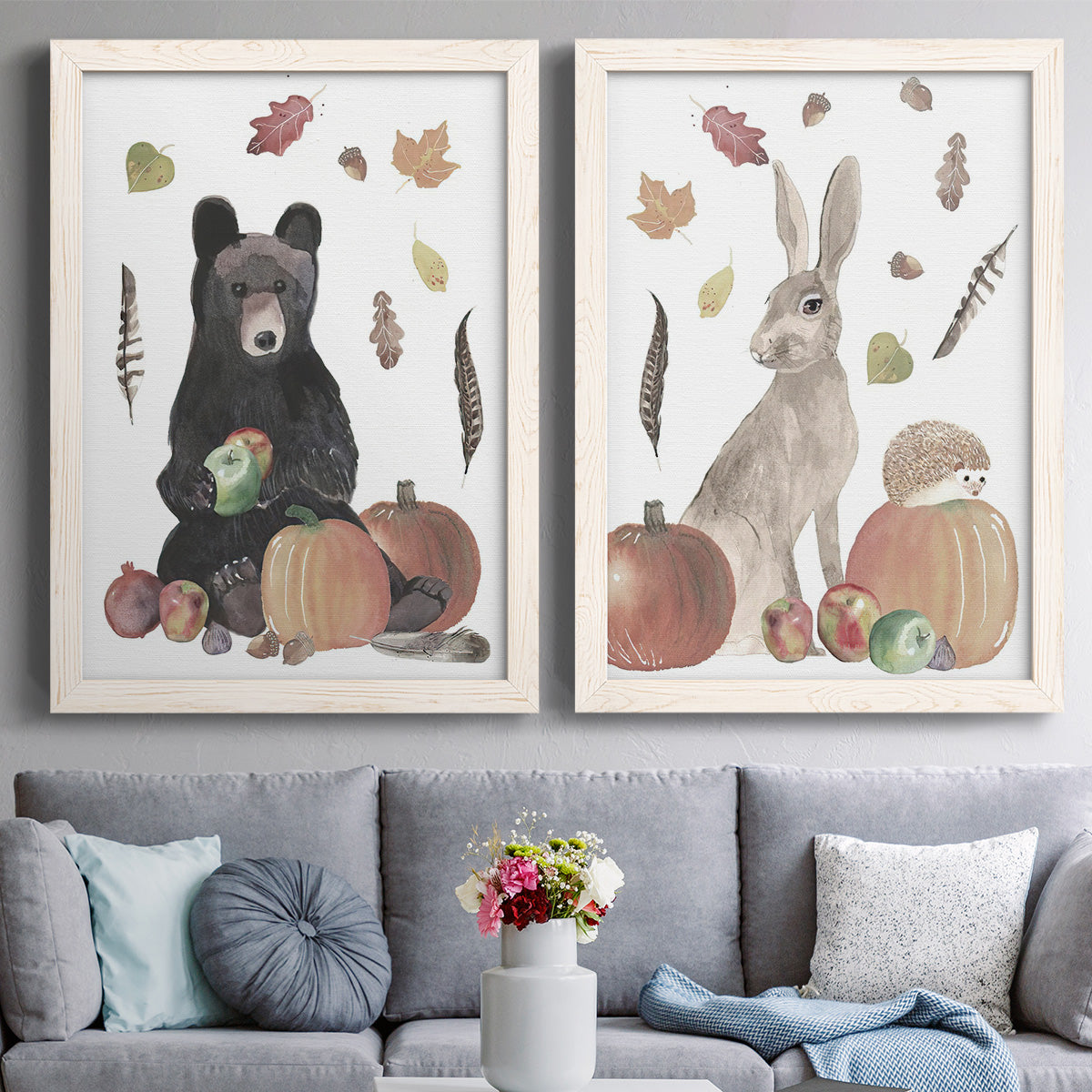 Set of two white barnwood framed canvas prints depicting playful watercolor wildlife surrounded by pumpkins acorns apples feathers and muted fall colors above a sofa in a contemporary living room.