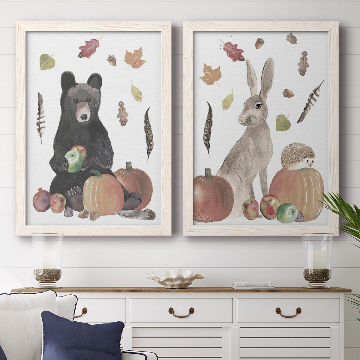 Set of two white barnwood framed canvas prints depicting two whimsical autumn illustrations of forest animals gathered among pumpkins apples feathers and seasonal foliage above a dining room sideboard.