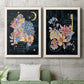 Set of two white barnwood framed canvas prints depicting a coordinated art pair of fantastical bouquets and celestial symbols on black rendered as whimsical floral collages above a sofa in a modern living room.