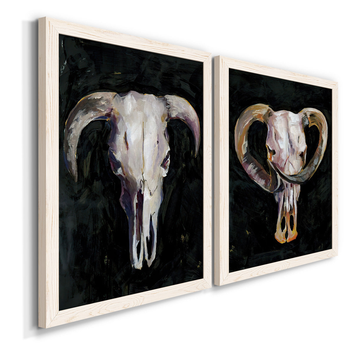 Angled view of a set of two white barnwood framed canvas prints featuring two dramatic Western animal paintings featuring ram and cattle skulls against black grounds.