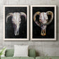 Set of two white barnwood framed canvas prints depicting a coordinated art pair of ram and cattle skulls against black grounds rendered as dramatic Western animal paintings above a sofa in a modern living room.