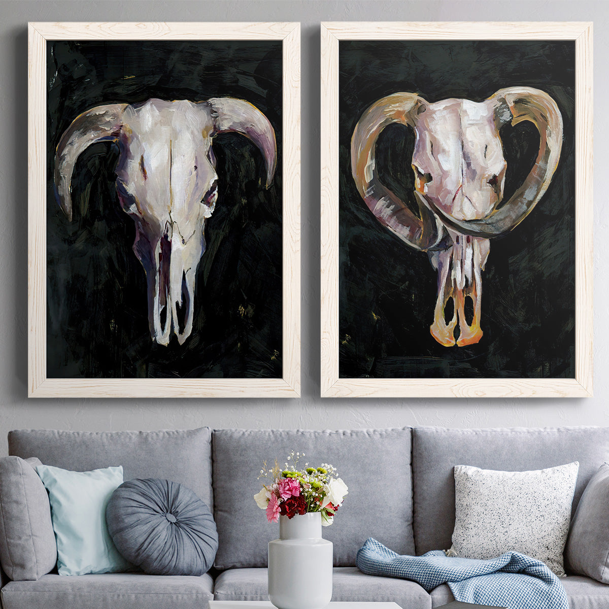 Set of two white barnwood framed canvas prints depicting paired dramatic Western animal paintings with curved horns, bone-white forms, and expressive dark brushwork above a sofa in a contemporary living room.