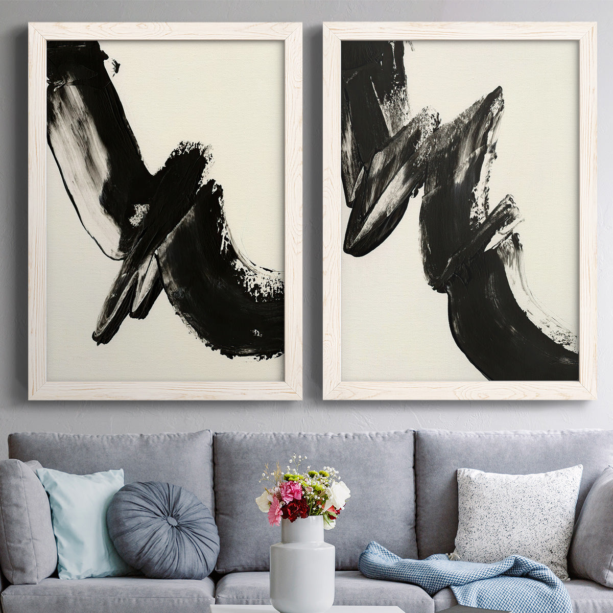 Set of two white barnwood framed canvas prints depicting paired bold gestural abstracts with thick ink-like marks, feathered edges, and cream negative space above a sofa in a contemporary living room.