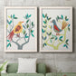 Set of two white barnwood framed canvas prints depicting a coordinated art pair of stylized colorful birds perched among leafy branches rendered as modern folk-art bird illustrations above a sofa in a modern living room.