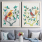 Set of two white barnwood framed canvas prints depicting paired modern folk-art bird illustrations with red yellow blue plumage, simplified leaves, and playful flat shapes above a sofa in a contemporary living room.