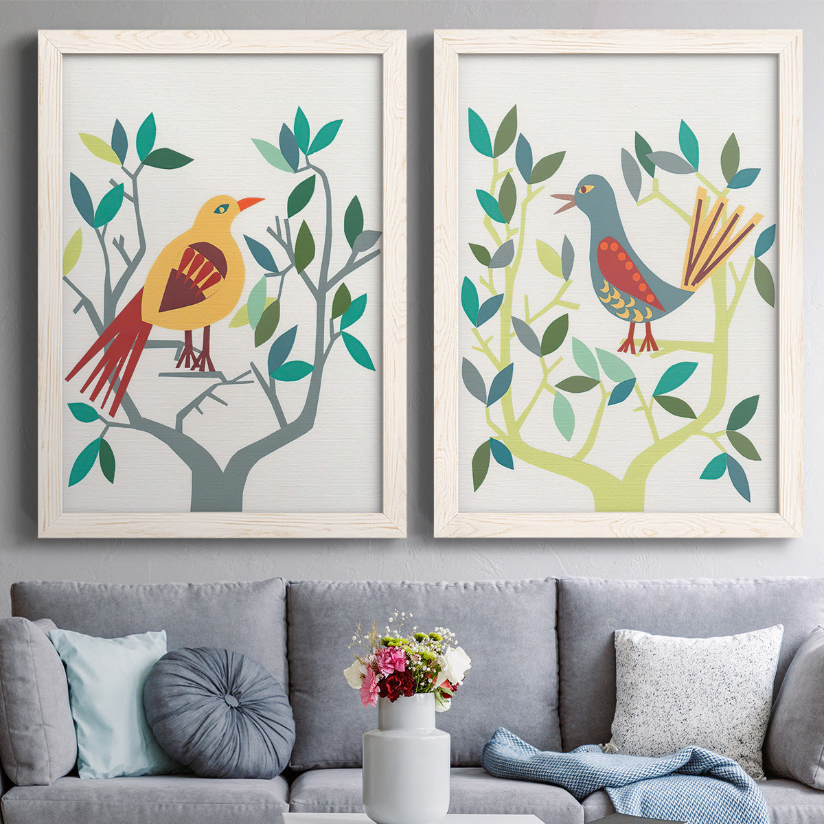 Set of two white barnwood framed canvas prints depicting paired modern folk-art bird illustrations with red yellow blue plumage, simplified leaves, and playful flat shapes above a sofa in a contemporary living room.