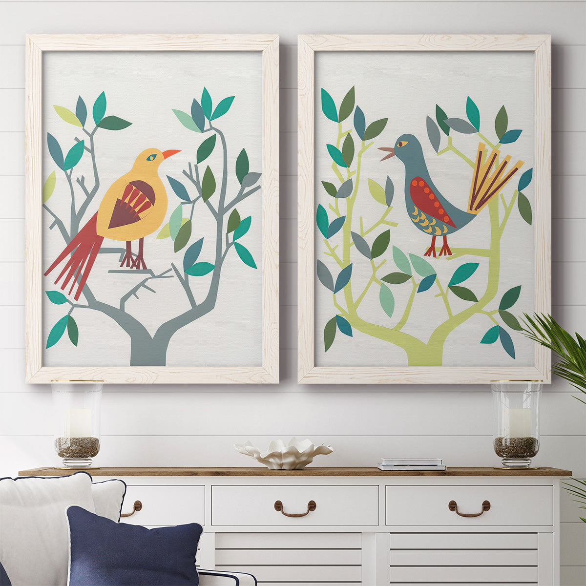 Set of two white barnwood framed canvas prints depicting a coordinated art pair of stylized colorful birds perched among leafy branches rendered as modern folk-art bird illustrations above a dining room sideboard.