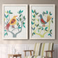 Set of two white barnwood framed canvas prints depicting two modern folk-art bird illustrations featuring stylized colorful birds perched among leafy branches above a sideboard in a cozy sitting room.