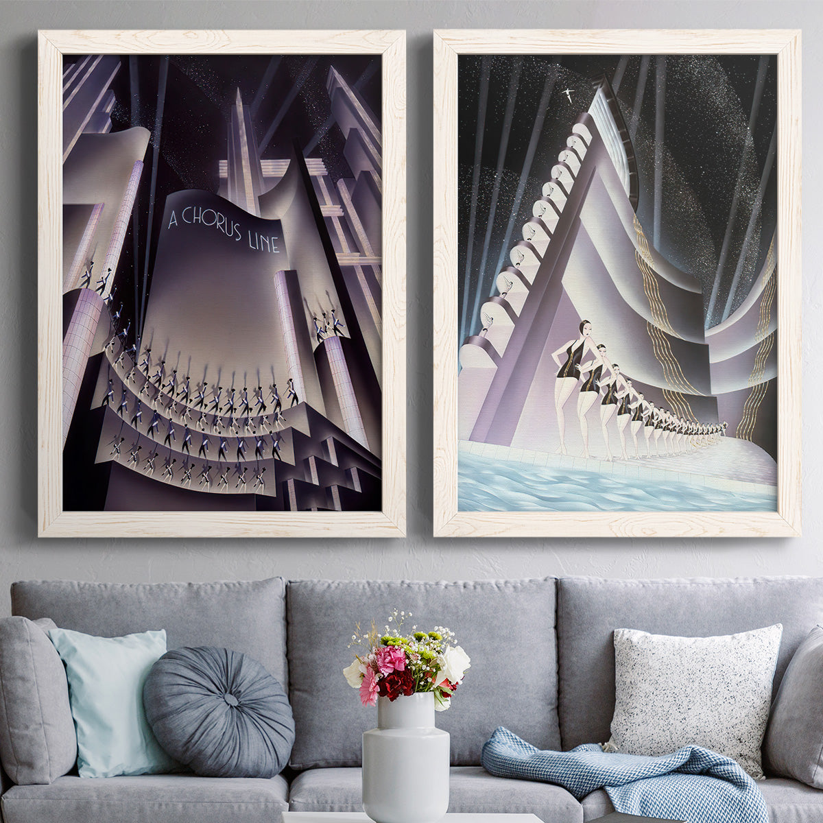 Set of two white barnwood framed canvas prints depicting paired vintage architectural illustrations with monumental skyscrapers, sweeping stairways, spotlights, and elegant nighttime figures above a sofa in a contemporary living room.