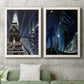 Set of two white barnwood framed canvas prints depicting a vintage urban pair of towering architecture searchlights stars and shadowed people at night above a sofa in a modern living room.