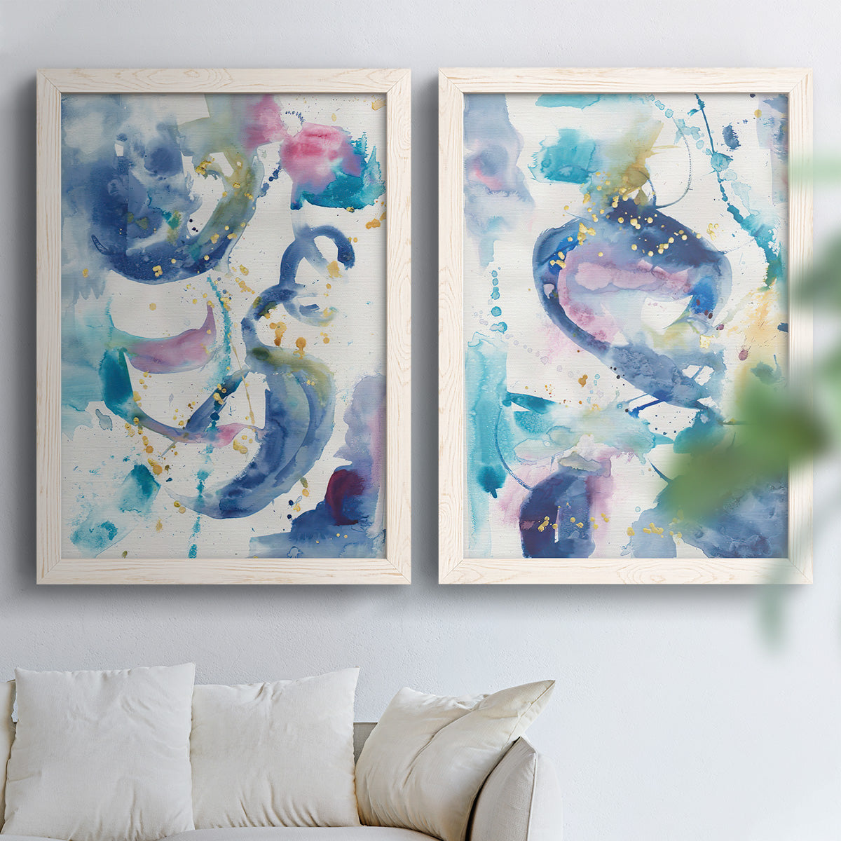Set of two white barnwood framed canvas prints depicting looping brush marks, splatters, and translucent overlapping washes arranged across two complementary compositions above a sofa in a bright living space.