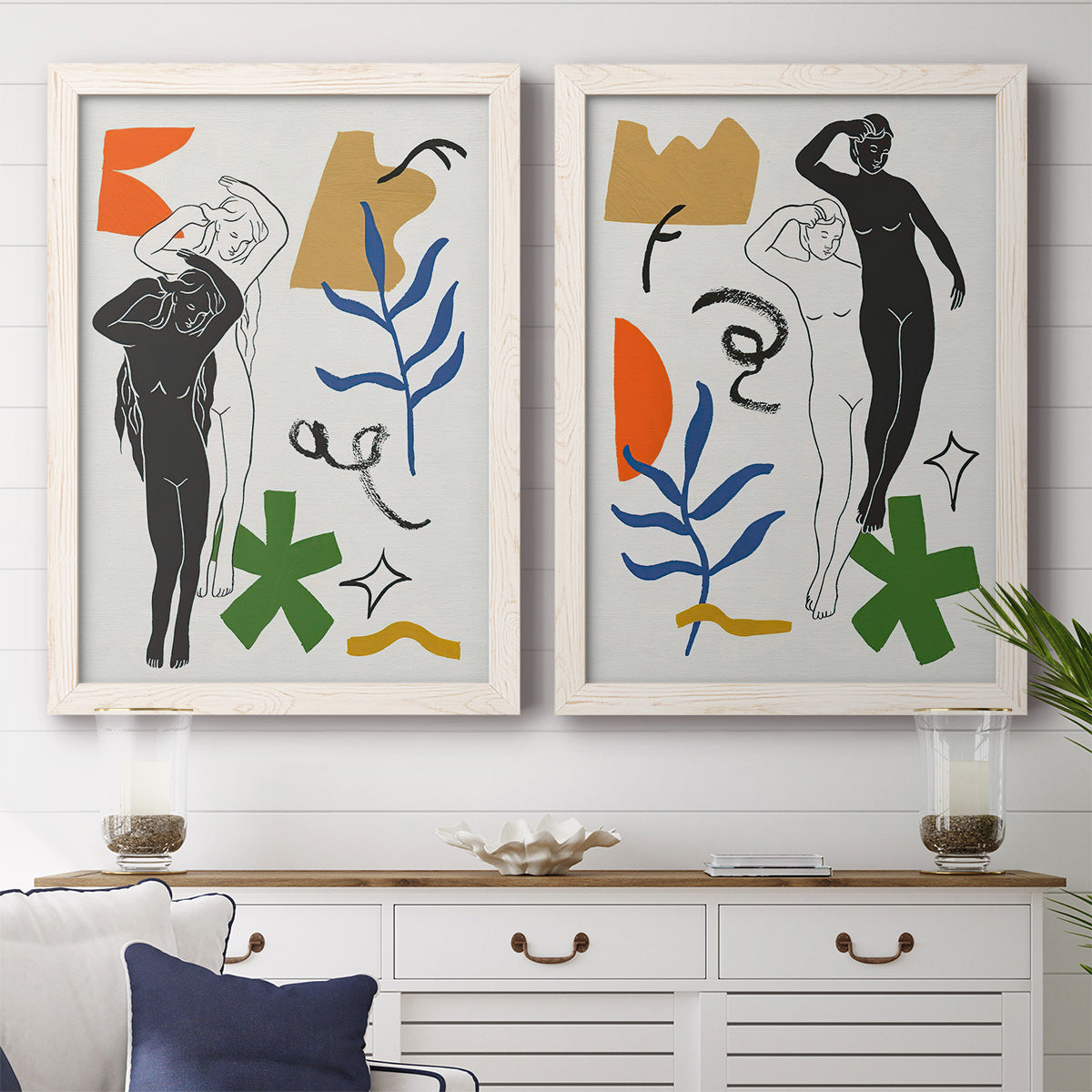 Set of two white barnwood framed canvas prints depicting stylized female figures surrounded by blue leaves green starbursts orange forms and gestural marks above a dining room sideboard.