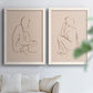 Set of two white barnwood framed canvas prints depicting warm brown contours, relaxed poses, and blush cream grounds arranged across two complementary compositions above a sofa in a bright living space.