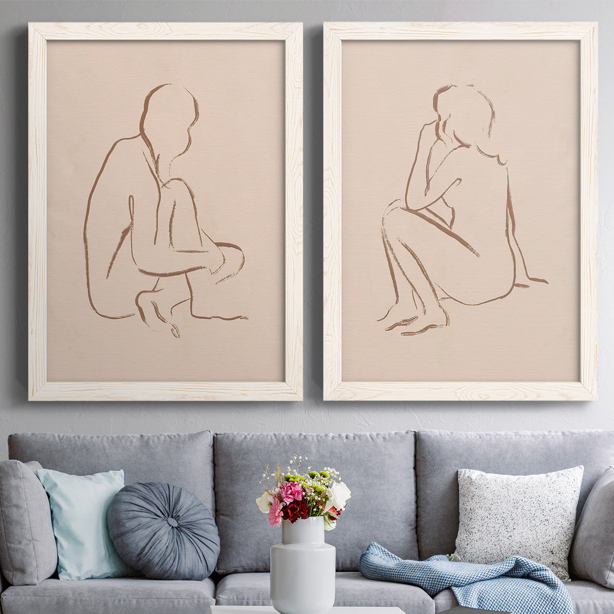 Set of two white barnwood framed canvas prints depicting paired minimalist figurative sketches with warm brown contours, relaxed poses, and blush cream grounds above a sofa in a contemporary living room.