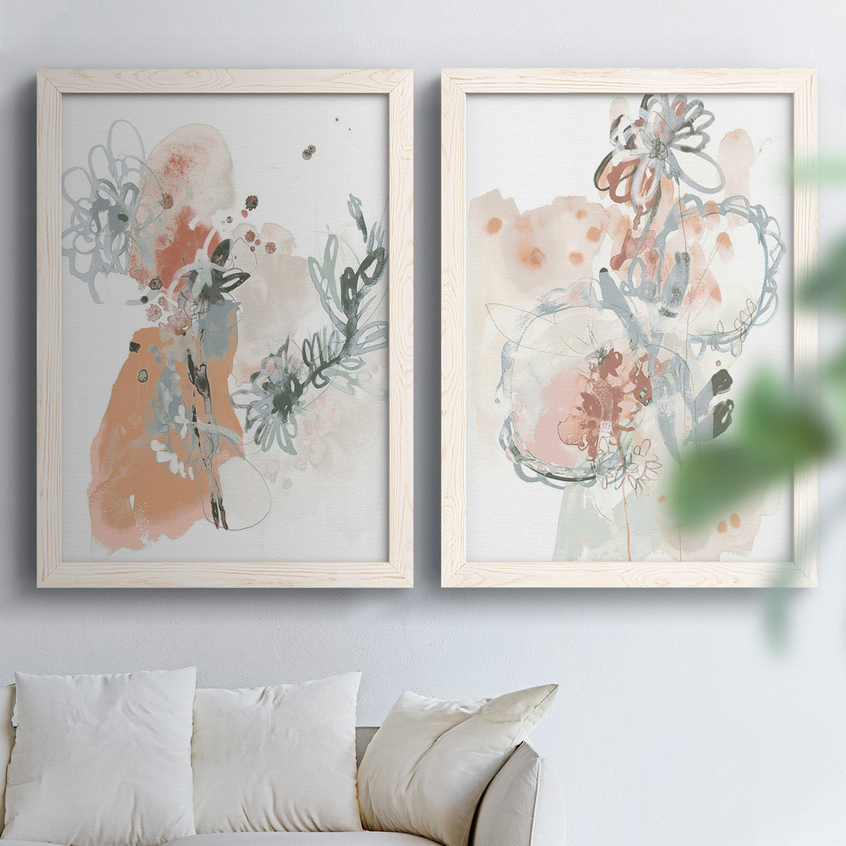Set of two white barnwood framed canvas prints depicting scribbled petals, translucent washes, and drifting stems arranged across two complementary compositions above a sofa in a bright living space.