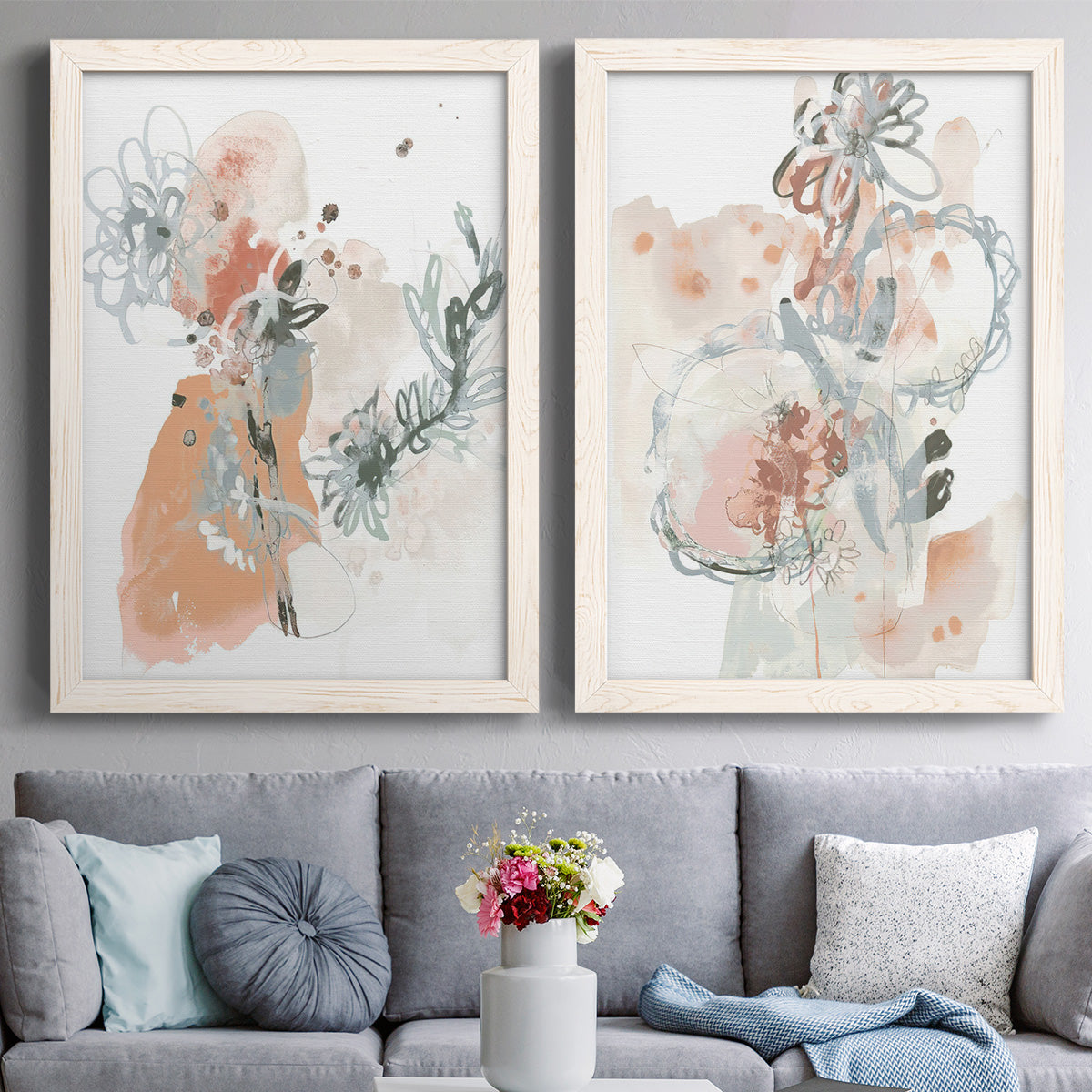 Set of two white barnwood framed canvas prints depicting paired abstract watercolor botanicals with scribbled petals, translucent washes, and drifting stems above a sofa in a contemporary living room.