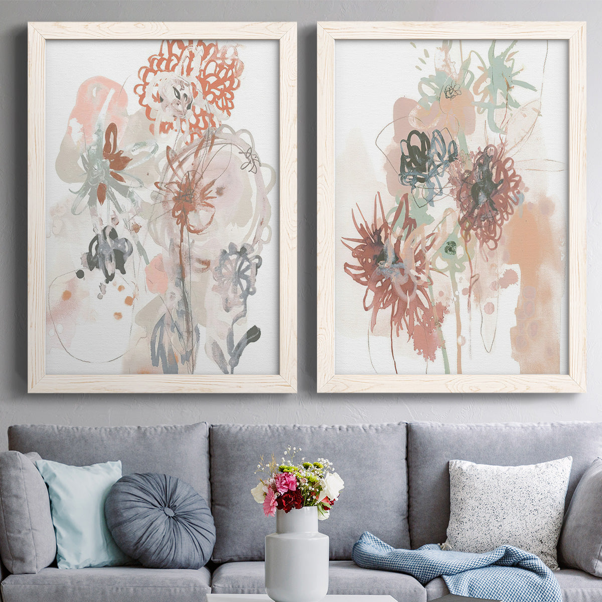 Set of two white barnwood framed canvas prints depicting paired abstract watercolor botanicals with scribbled petals, translucent washes, and drifting stems above a sofa in a contemporary living room.
