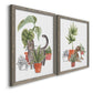 Angled view of a set of two gray barnwood framed canvas prints featuring two whimsical botanical animal illustrations featuring cats tucked among collections of potted houseplants.
