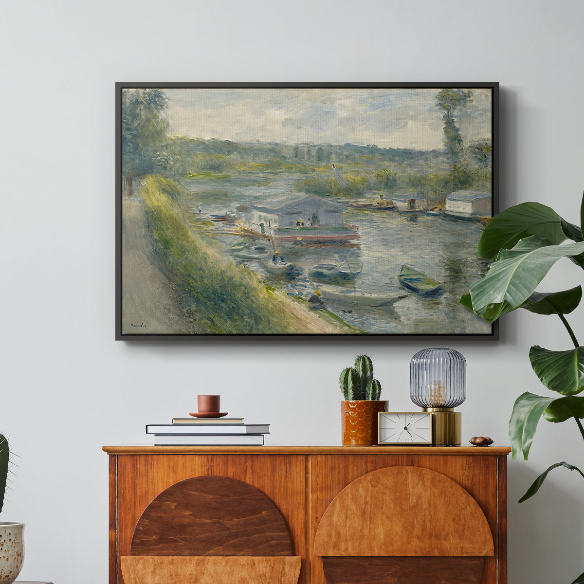 Wash-House Boat at Bas-Meudon, c. 1874 by Pierre-Auguste Renoir in a black floater framed canvas featuring a floating wash-house boat along a riverbank with figures working nearby, displayed above a wood console cabinet.