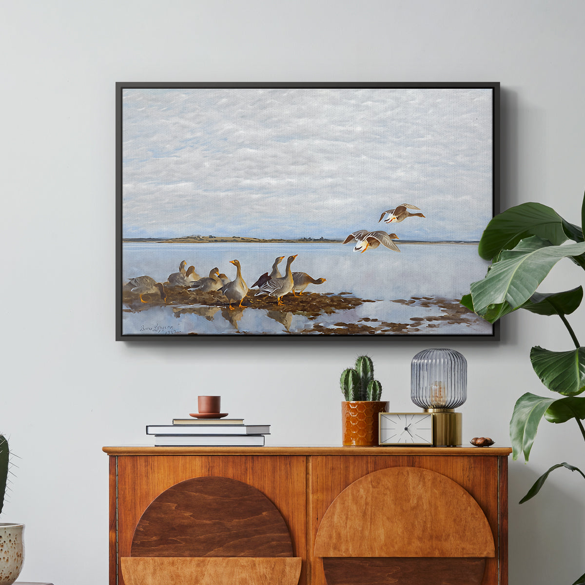 Greylag Geese (Grågäss), 1922 by Bruno Liljefors in a black floater framed canvas featuring a group of geese gathered near water in a natural wetland landscape, displayed above a wood console cabinet.