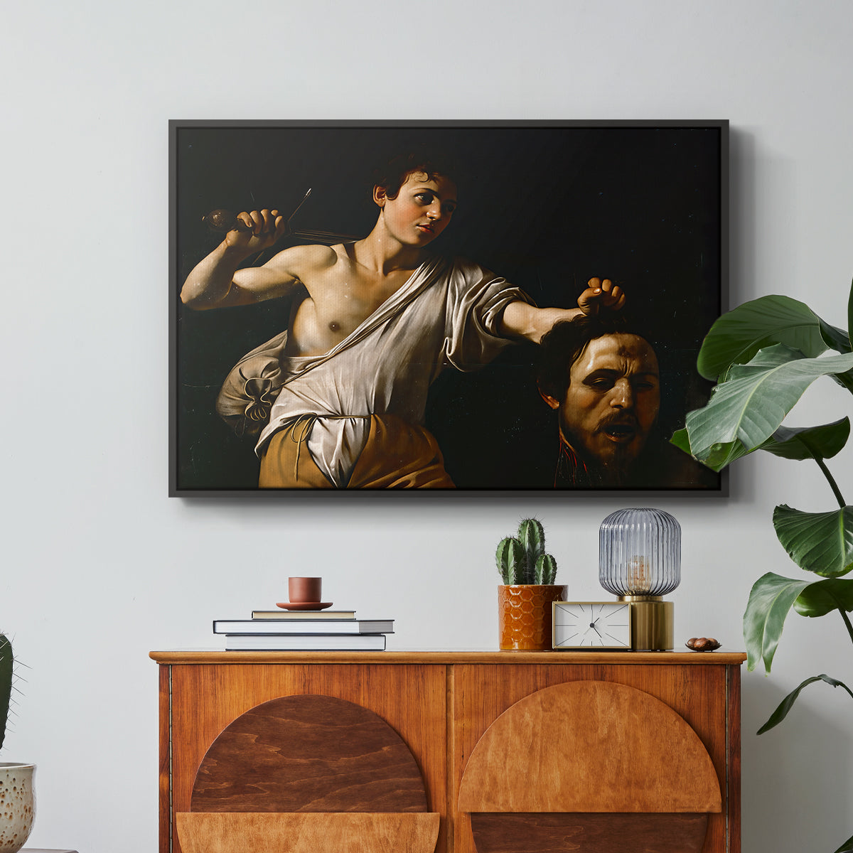 David with the Head of Goliath (Vienna Version), c. 1607 by Caravaggio in a black floater framed canvas featuring a young David holding the severed head of Goliath against a dark background, displayed above a wood console cabinet.
