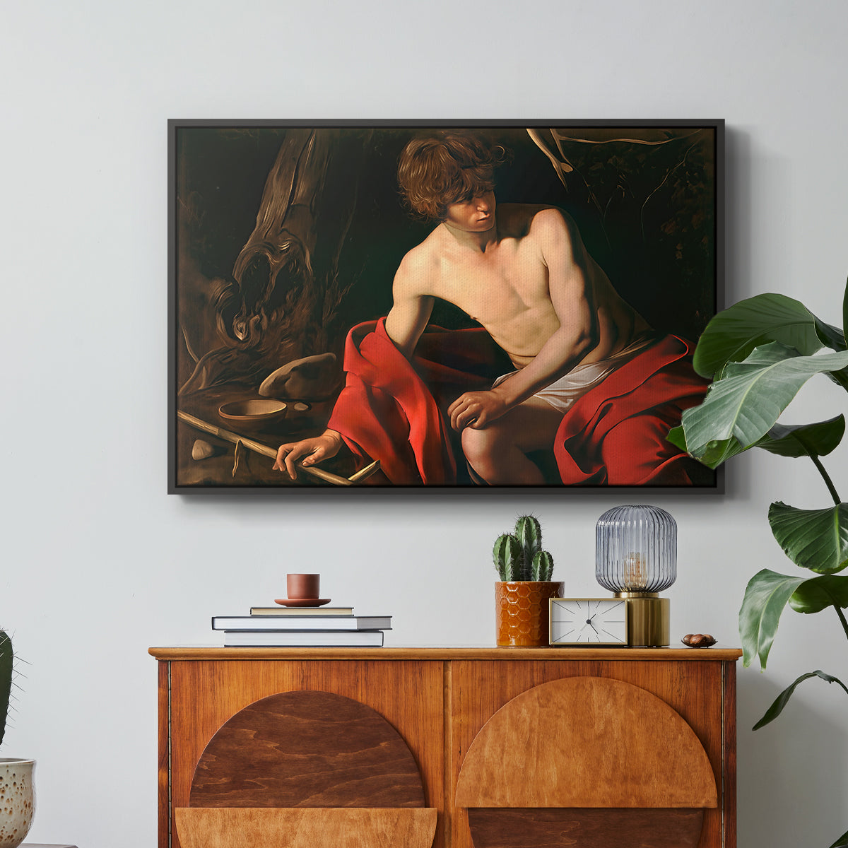 Saint John the Baptist in the Wilderness, c. 1603-1604 by Caravaggio in a black floater framed canvas featuring a young Saint John seated in wilderness with staff and red drapery, displayed above a wood console cabinet.