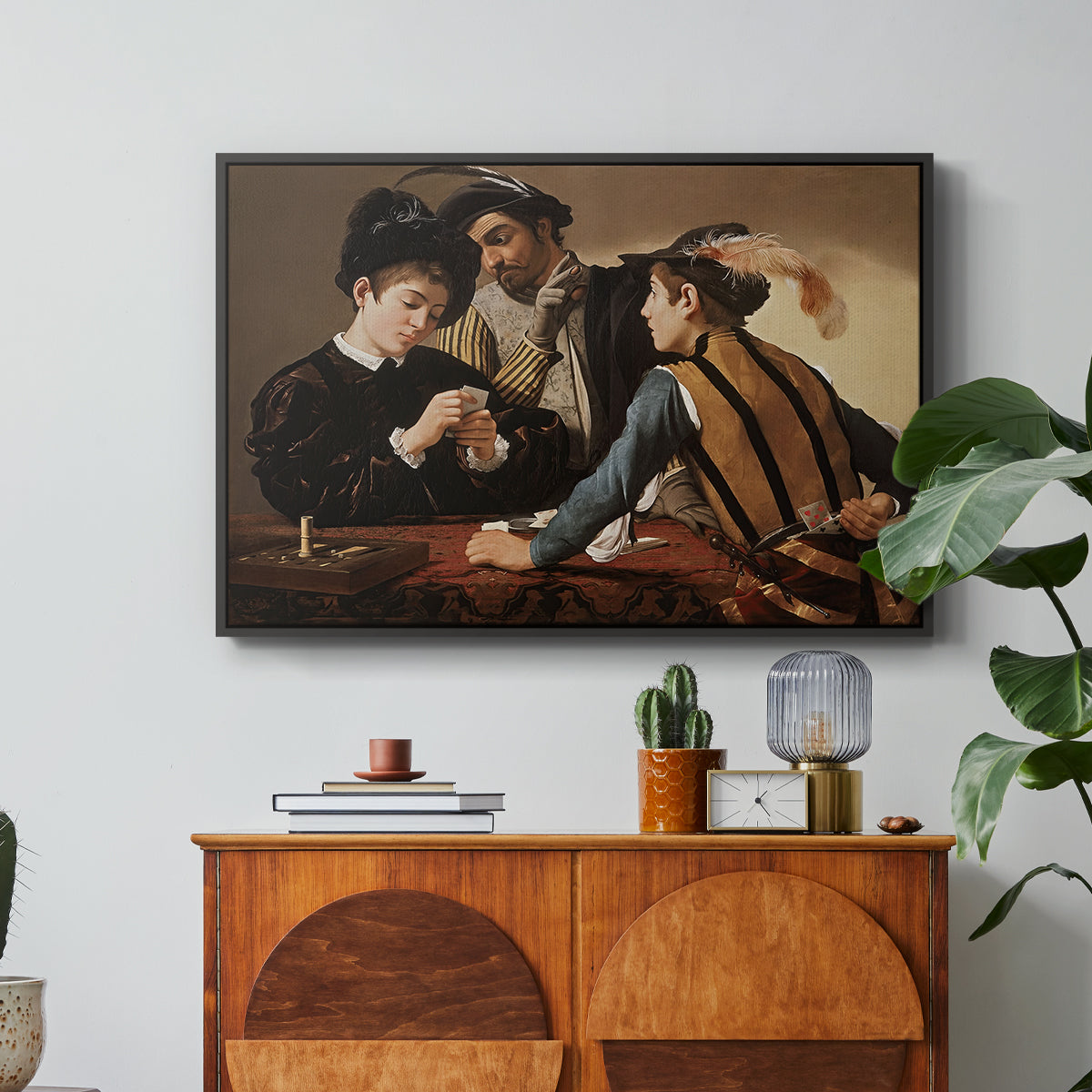The Cardsharps, c. 1594 by Caravaggio in a black floater framed canvas featuring two men cheating at cards while a third figure secretly observes behind them, displayed above a wood console cabinet.