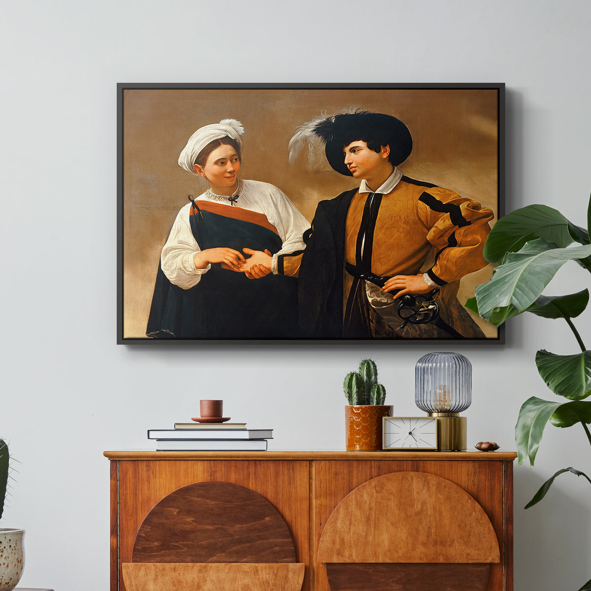 The Fortune Teller (Capitoline Version), c. 1599 by Caravaggio in a black floater framed canvas featuring a woman reading man's palm while discreetly removing his ring, displayed above a wood console cabinet.
