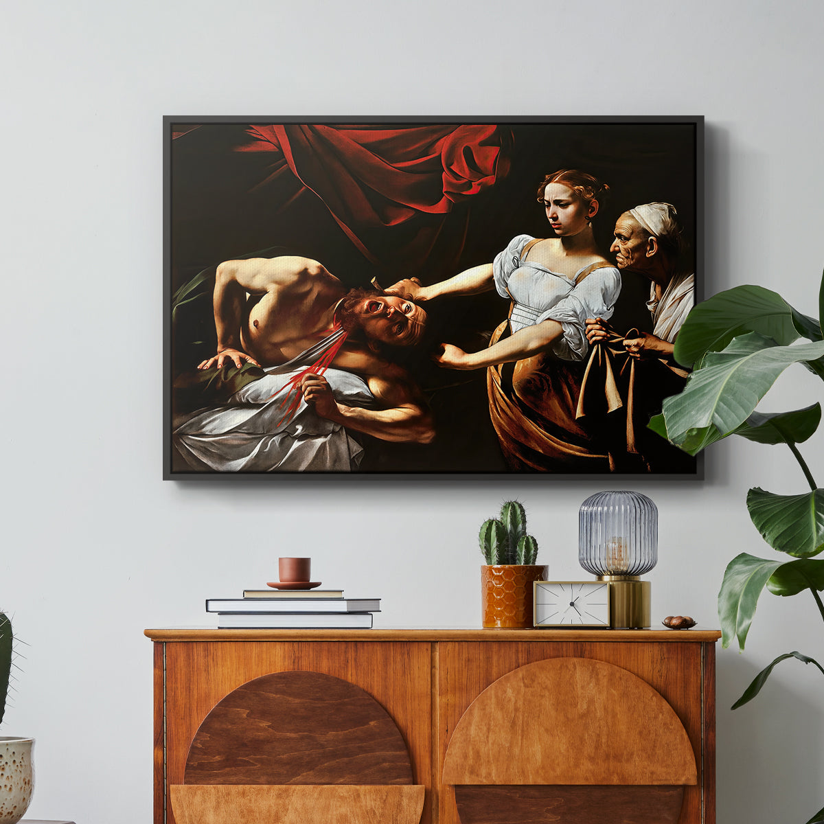 Judith Beheading Holofernes, c. 1599 by Caravaggio in a black floater framed canvas featuring Judith cutting off Holofernes' head as maidservant assists beside her, displayed above a wood console cabinet.