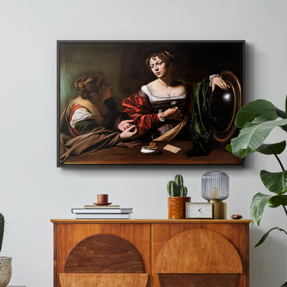 Martha and Mary Magdalene, c. 1598 by Caravaggio in a black floater framed canvas featuring a two women seated together in conversation with symbolic objects on table, displayed above a wood console cabinet.