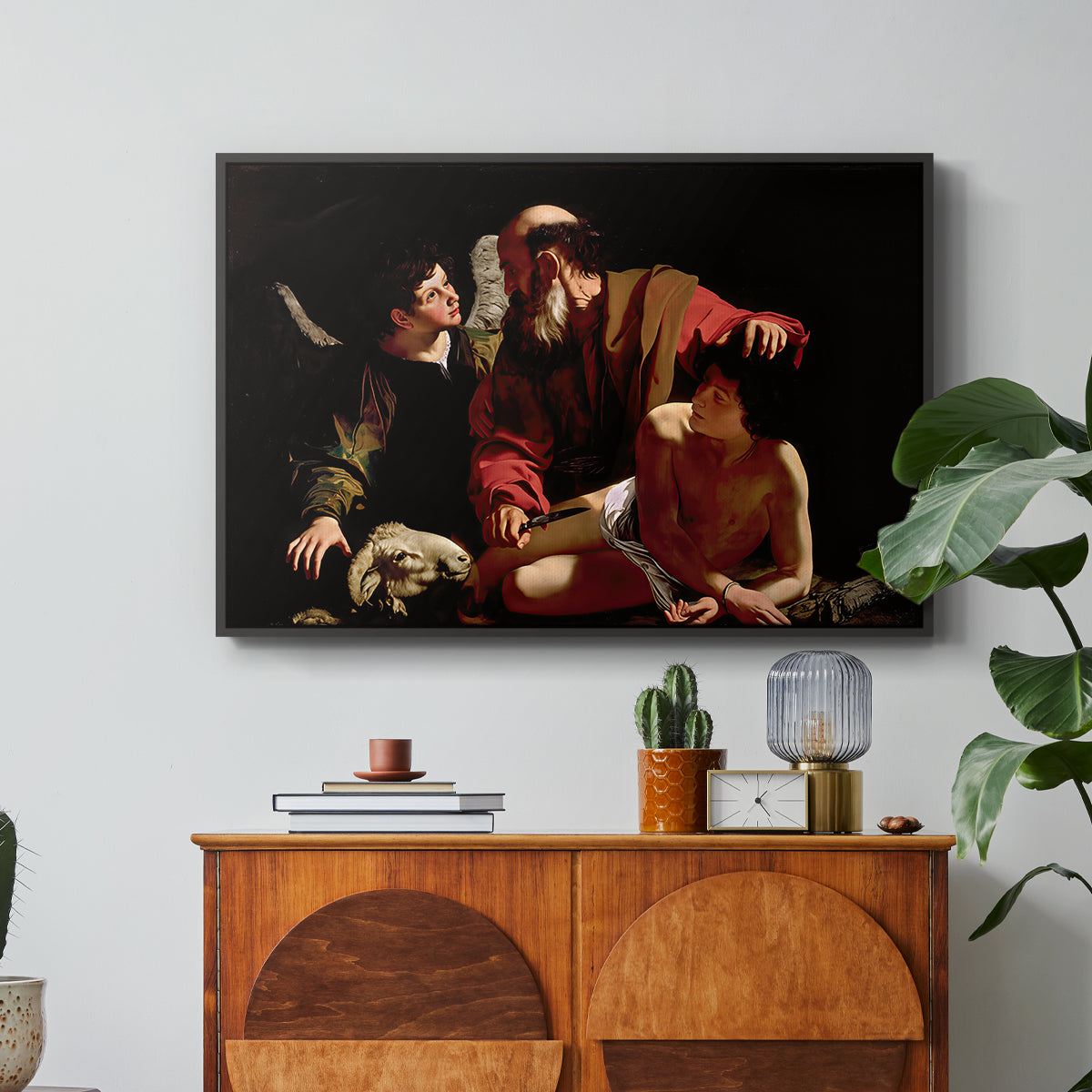 The Sacrifice of Isaac (Uffizi Version), c. 1603 by Caravaggio in a black floater framed canvas featuring Abraham holding Isaac while angel intervenes to stop the sacrifice, displayed above a wood console cabinet.