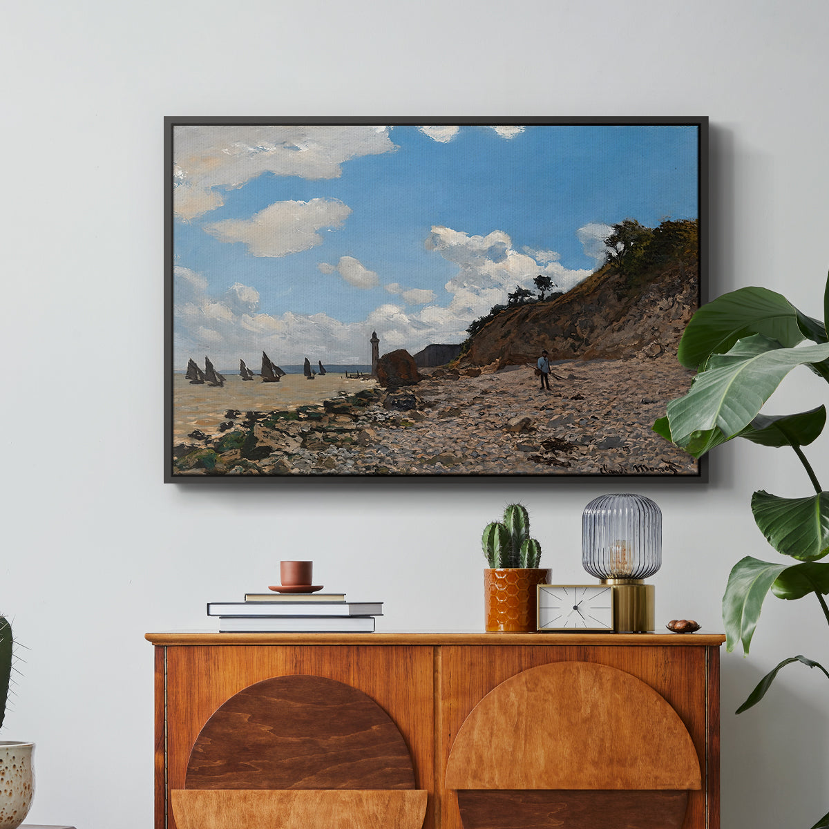 The Beach at Honfleur, 1864 by Claude Monet in a black floater framed canvas featuring a coastal beach scene with figures gathered along shoreline and boats nearby, displayed above a wood console cabinet.