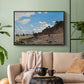 Black floater framed canvas featuring a coastal landscape with boats and people along shore, displayed above a sofa in a modern living room.