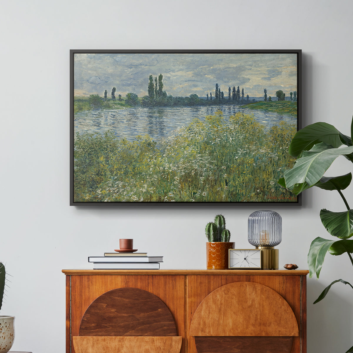 Banks of the Seine, Vétheuil, 1880 by Claude Monet in a black floater framed canvas featuring a riverbank landscape with water, trees, and distant village buildings, displayed above a wood console cabinet.