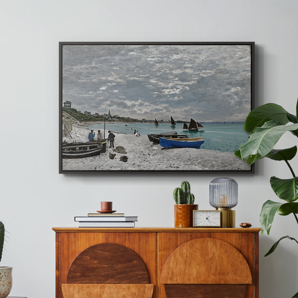 The Beach at Sainte-Adresse, 1867 by Claude Monet in a black floater framed canvas featuring a seaside promenade with figures, flags, and boats along the coast, displayed above a wood console cabinet.