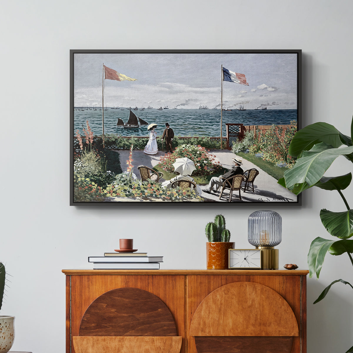 Garden at Sainte-Adresse, 1867 by Claude Monet in a black floater framed canvas featuring a seaside garden with flowers, flags, and figures overlooking the water, displayed above a wood console cabinet.