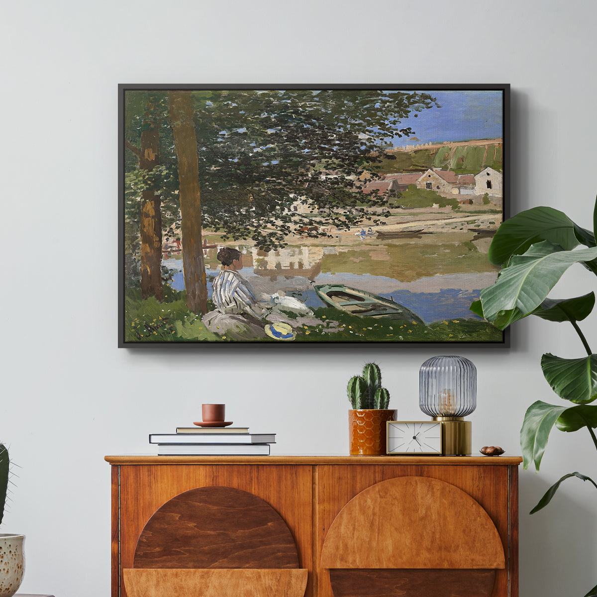 On the Bank of the Seine, Bennecourt, 1868 by Claude Monet in a black floater framed canvas featuring a woman and child seated along riverbank beneath trees in soft daylight, displayed above a wood console cabinet.