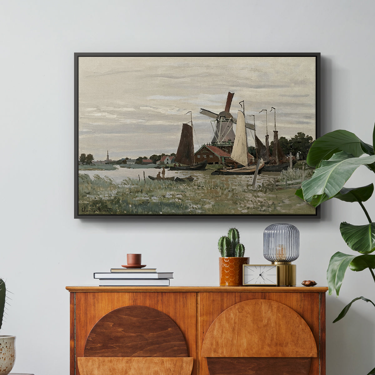 Windmill at Zaandam, 1871 by Claude Monet in a black floater framed canvas featuring a single windmill beside water with surrounding buildings and sky, displayed above a wood console cabinet.