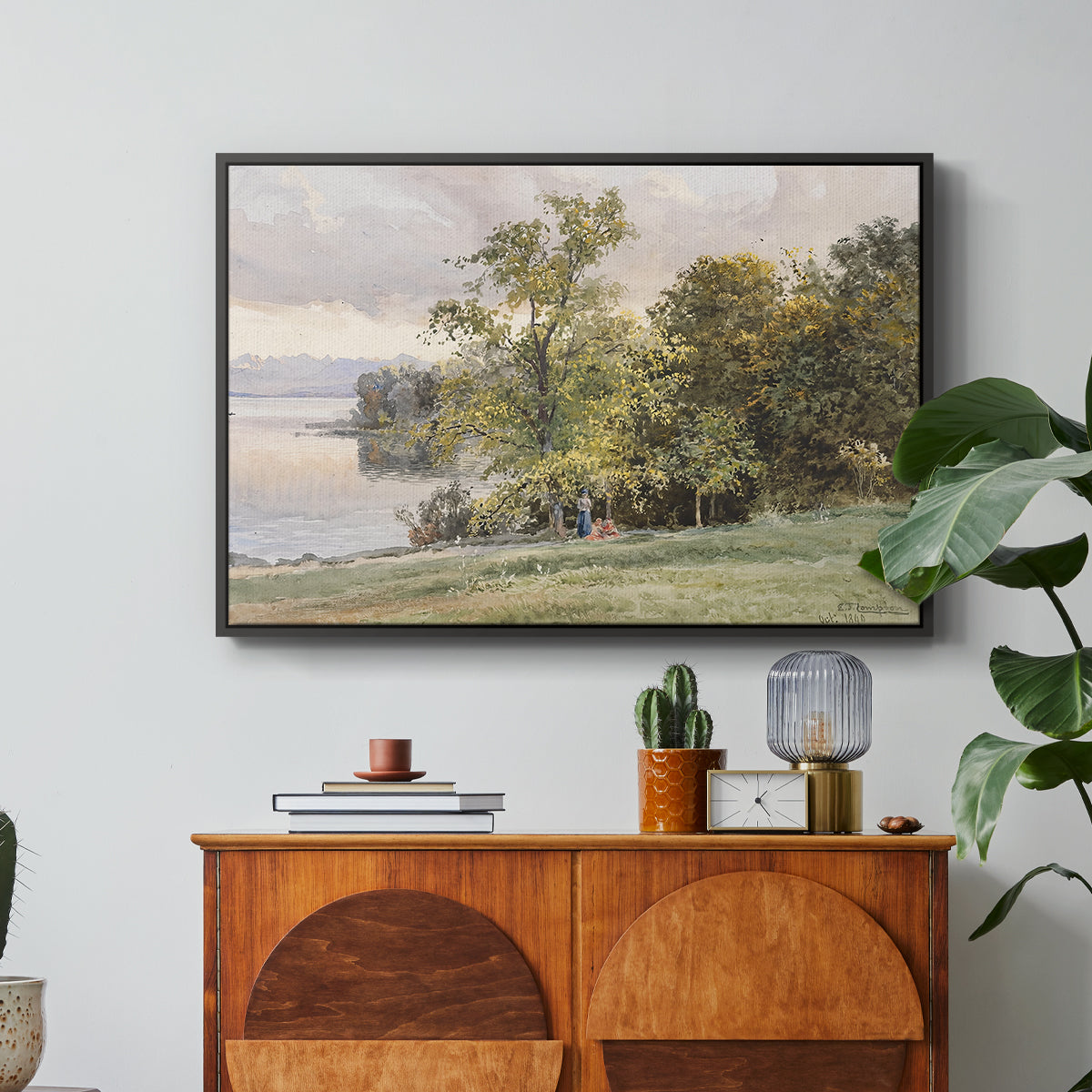 Landscape at Lake Starnberg, 1890 by Edward Theodore Compton in a black floater framed canvas featuring a lakeside landscape with calm water and trees along shoreline, displayed above a wood console cabinet.