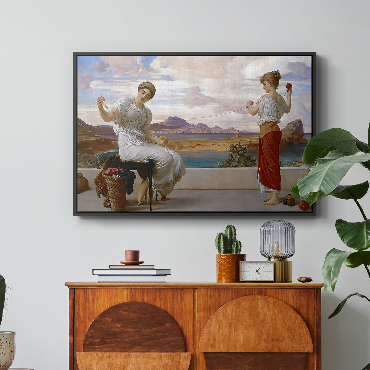 Winding the Skein, c. 1878 by Frederic Leighton in a black floater framed canvas featuring a woman holding thread while another figure helps wind yarn in classical setting, displayed above a wood console cabinet.