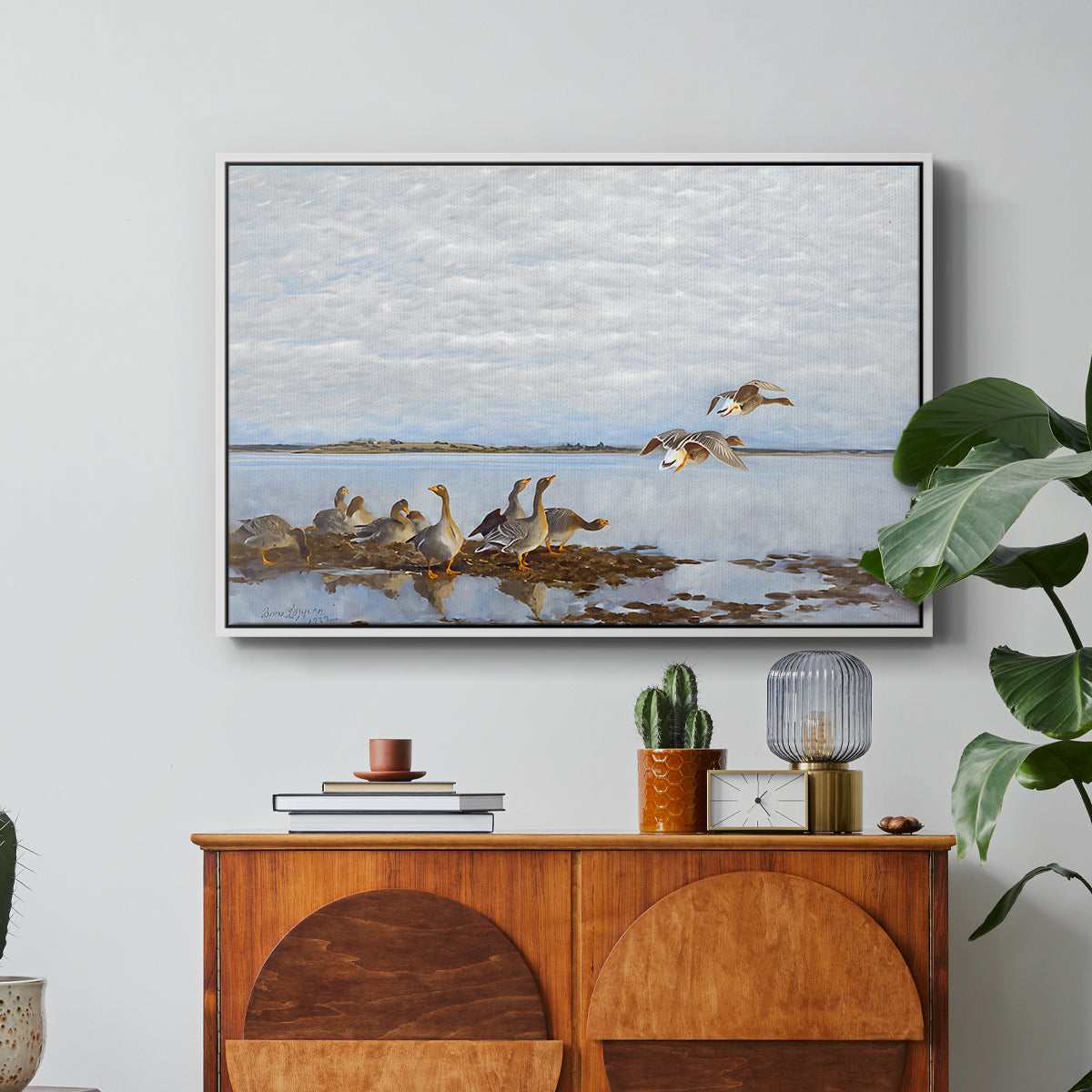 Greylag Geese (Grågäss), 1922 by Bruno Liljefors in a white floater framed canvas featuring a group of geese gathered near water in a natural wetland landscape, displayed above a wood console cabinet.